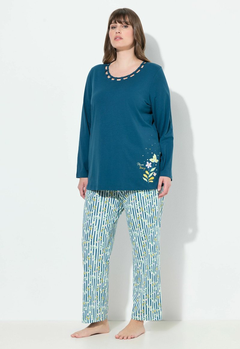 Kvinde iført teal langærmet top med blomsterprint og stribede blomsterpyjamasbukser, stående barfodet mod hvid baggrund.
