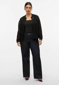 Veste bomber en dentelle noire avec fermeture par bouton, assortie à un haut noir ajusté et un jean taille haute en denim bleu foncé. Présente des coutures audacieuses et des chaussures à bout pointu.