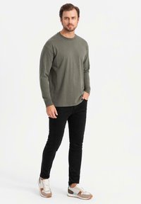 Olivgrön långärmad skjorta, svarta slim-fit byxor och grå sneakers med bruna och blå detaljer, stående mot en vit bakgrund.