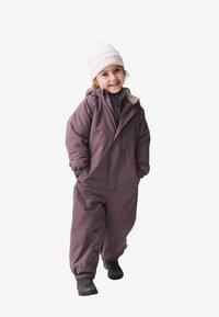 Lila isolierter Schneeanzug mit Reißverschluss vorne, elastischen Bündchen und einem beigen Kragen. Genossen über einem grauen Oberteil, kombiniert mit schwarzen Stiefeln und einer weißen Mütze.