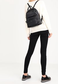 Musta nailonireppu, jossa on kaksi ulkoista vetoketjupussia, säädettävät hihnat ja sileä pinta. Mallilla on päällä valkoinen neule ja mustat legginsit.