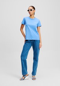 Világoskék pamut póló rövid ujjakkal, közepes mosású farmernadrággal párosítva. A modellen fehér napszemüveg és világoskék szandál van.