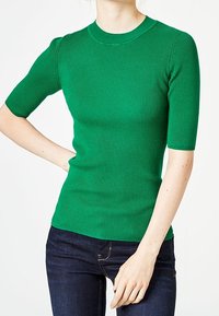 Zelený žebrovaný top s krátkým rukávem a kulatým výstřihem. Má přiléhavý střih a texturovanou tkaninu. Nosený s tmavými džínami.