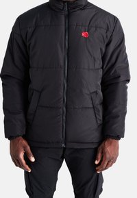 Mann in schwarzer gesteppter Jacke mit rotem Rosenemblem auf der Brust, Reißverschluss vorne, Seitentaschen und schwarzen Hosen.