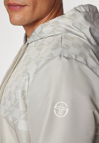 Chaqueta con capucha de color gris claro que presenta un patrón de diamantes en los hombros y un logo circular en la manga, con un acabado liso y texturizado.