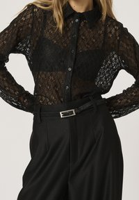 Blusa di pizzo nero con chiusura a bottoni, tessuto trasparente e colletto, abbinata a pantaloni neri a vita alta e una cintura sottile.