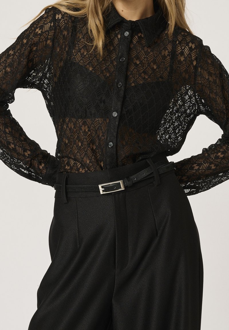 Blusa di pizzo nero con chiusura a bottoni, tessuto trasparente e colletto, abbinata a pantaloni neri a vita alta e una cintura sottile.