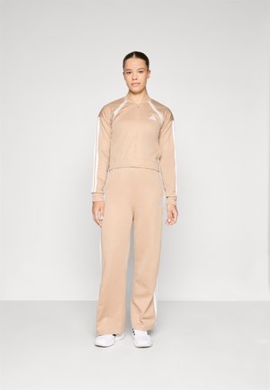 Veste zippée athlétique beige avec des rayures blanches et un pantalon assorti. Tissu doux, coupe décontract�ée, design minimaliste, avec une marque discrète.