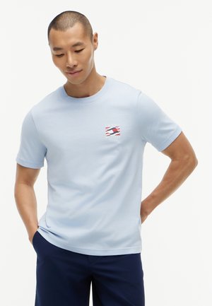 Νέος άνδρας που φοράει ανοιχτό μπλε μπλουζάκι Tommy Hilfiger με λογότυπο στο στήθος και σκούρο μπλε παντελόνι, στέκεται με τα χέρια στις τσέπες.