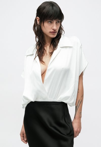 Žena s tamnom valovitom kosom nosi labavu bijelu bluzu s dubokim V-izrezom i crnu suknju, pokazujući tetovaže na desnoj podlaktici.