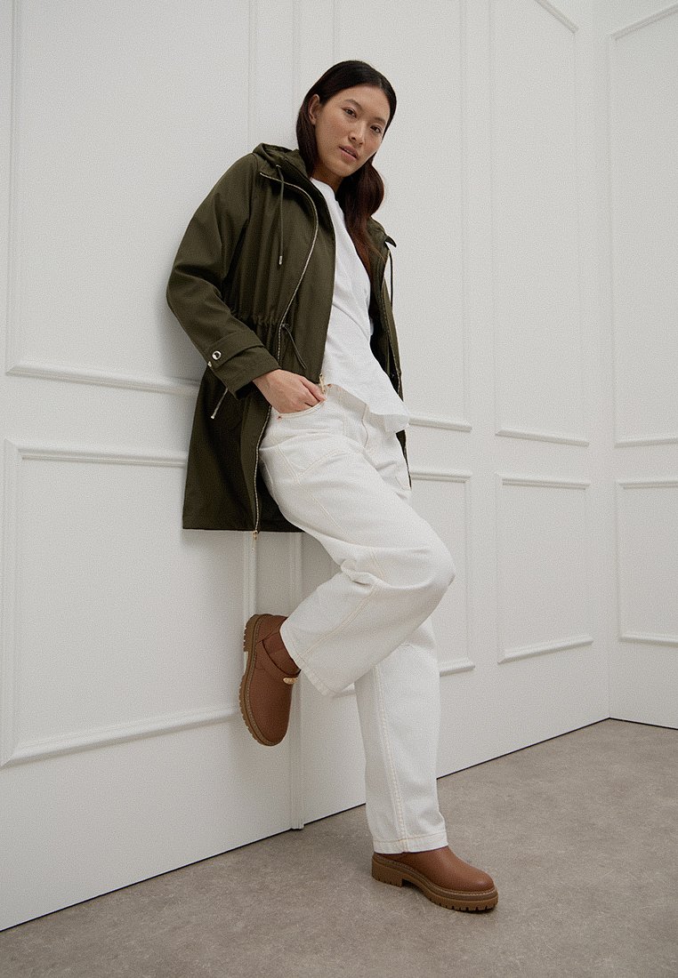 Vrouw leunend tegen een witte paneelwand, gekleed in een groene capuchonjack, een wit shirt en broek, en bruine enkellaarsjes.