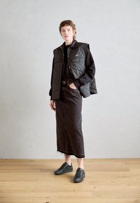 Gilet imbottito nero sopra un maglione nero, abbinato a una lunga gonna di jeans nera e sneakers grigio scuro. La gonna ha un orlo sfrangiato.