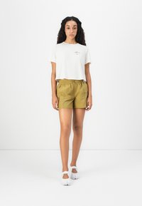 T-shirt blanche raccourcie avec un petit graphique, shorts verts olive avec une taille élastique, et chaussures blanches avec des brides de cheville. Design simple et décontracté.