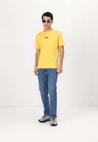 Tricou galben din bumbac cu un mic logo, blugi albaștri și adidași gri cu accente negre. Model purtând ochelari de soare, stând pe un fundal alb.