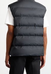 Svart pufferväst med hög krage, quiltat mönster och horisontella sömmar. Tillverkad av ett slätt, glänsande material. Bärs över en vit skjorta.