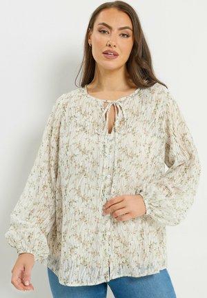 Donna che indossa una camicetta leggera a maniche lunghe con motivo floreale, bottoni e collo annodato, abbinata a jeans blu, in piedi davanti a uno sfondo bianco.