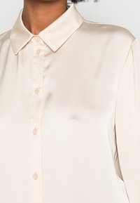 Blouse en satin couleur crème avec un col pointe, fermeture à boutons sur le devant et texture lisse avec une coupe ajustée.