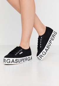 Black canvas platform sneakers with prominent white "SUPERGA" branding along the sole, featuring round laces and a rounded toe design.