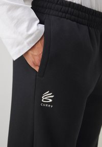 Pantalones de chándal negros con cinturilla elástica y bolsillos laterales. Presenta un logo bordado en blanco "Curry" en la zona del bolsillo izquierdo.
