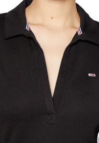 Black polo shirt with a collar, featuring a partial button placket, and a small logo patch with red, white, and blue accents on the collar.