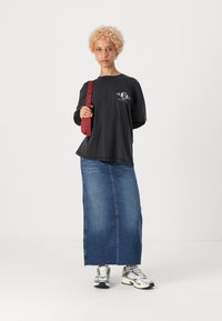 T-shirt noir oversize avec imprimé graphique, associé à une longue jupe en denim. Un sac à bandoulière rouge et des baskets argentées complètent le look.