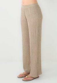 Beige, geplooide wijde broek met een gestructureerde afwerking en een elastische tailleband, gecombineerd met platte sandalen met een gouden accent.