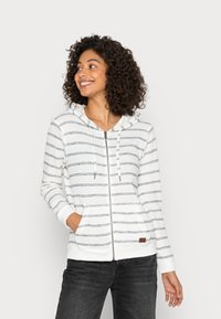 White zip-up hoodie with navy stripes, a drawstring hood, front pockets, and ribbed cuffs. Notable brown label on the hem.