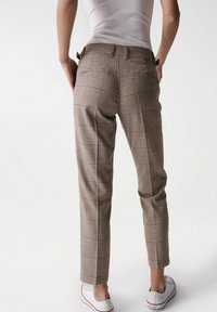 Personne portant un pantalon à carreaux taille haute et un débardeur blanc, se tenant avec les mains dans les poches et des baskets blanches sur un fond uni.