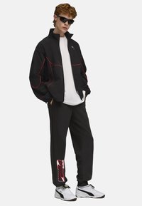 Sort træningsjakke med rød piping, parret med sorte sweatpants med logo-print. Hvid T-shirt og hvide sneakers fuldender looket.