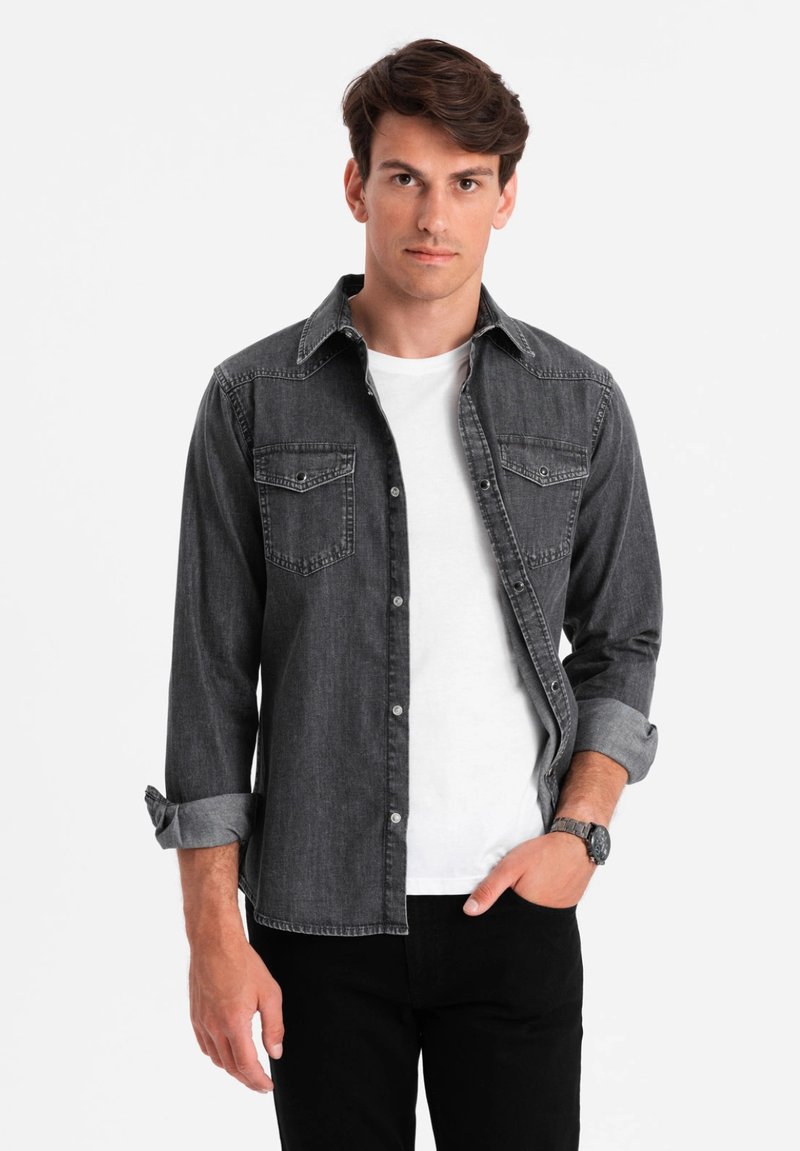 Grijs denimshirt met twee voorkruisen, button-down kraag en opgerolde mouwen over een wit T-shirt, gecombineerd met zwarte broek.