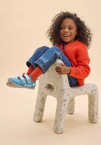 Un bambino è seduto su una sedia di colore grigio chiaro, con una texture particolare, realizzata in materiale riciclato. Indossa un maglione arancione brillante, scarpe blu con fibbie e pantaloni di jeans.