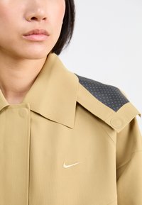 Veste beige avec un grand col, comportant un empiècement d'épaule à motif gris foncé et un petit logo brodé sur la poitrine.