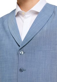 Helles blauer Blazer mit weicher Textur, ausgestattet mit einem Reverskragen, zwei Knöpfen und einer Brusttasche. Weiße Hemd mit Kragen ist darunter sichtbar.