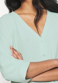 Haut vert menthe à col en V avec manches trois-quarts et coupe décontractée. Il présente un détail de bouton au niveau de l'encolure et une texture lisse et douce.