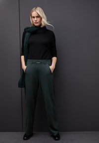 Suéter negro, pantalones de pierna ancha verde oscuro con bucle de cinturón texturizado y detalle de hebilla, combinados con una bufanda verde esponjosa. Zapatos negros.