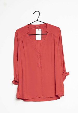 Hellrote Langarmbluse mit V-Ausschnitt, drei Knöpfen und hochgekrempelten Ärmeln, die zu Knoten gebunden sind, hängend an einem schwarzen Kleiderbügel mit weißem Preisschild.