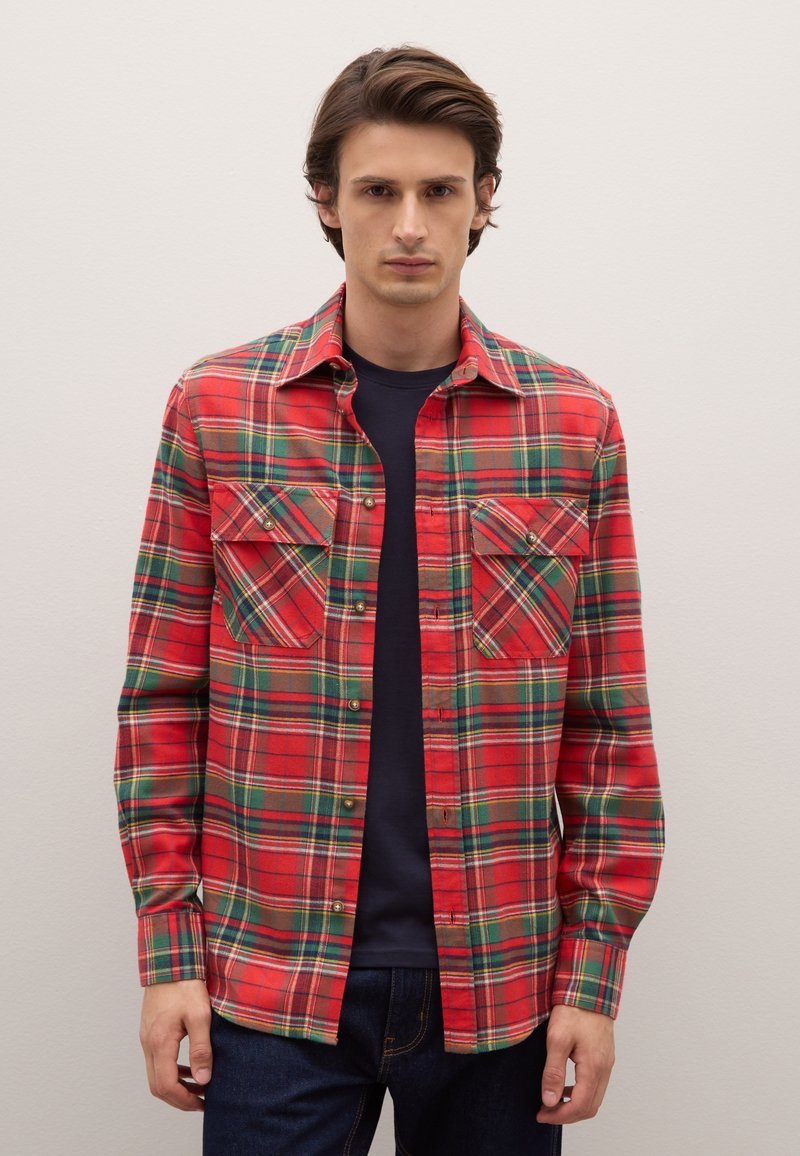 Camicia di flanella a quadri rossi con accenti verdi e gialli, dotata di due tasche frontali, chiusura con bottoni e maniche lunghe. Indossata sopra una maglia blu navy.