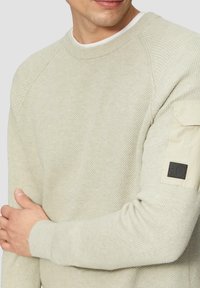 Jasno beżowy sweter o teksturowanym splocie, okrągłym dekolcie i ściągaczach na rękawach. Posiada materiałową naszywkę na lewym rękawie.