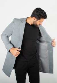 Blazer gris avec une fermeture à un bouton, des revers à cran et une doublure lisse et clair. Présente des lignes épurées et une coupe ajustée.