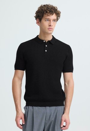 Junger Mann mit lockigem Haar, der ein schwarzes, kurzärmliges Strick-Poloshirt und graue, plissierte Hosen trägt und vor einem einfarbigen Hintergrund steht.