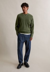 Pulover verde măsliniu, cu guler rotund, din material texturat; asortat cu blugi largi din denim închis la culoare și mocasini negri. Colier simplu din argint.