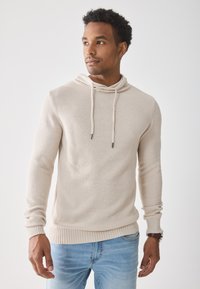 Haut à capuche en tricot beige clair avec un motif texturé, col à cordon, manches longues et poignets côtelés. Porté par-dessus un jean bleu.