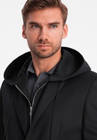 Schwarze Kapuzenjacke mit Reißverschluss, über einem dunkelgrauen Poloshirt getragen. Hat eine glatte Textur und ein minimalistisches Design.