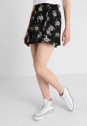 Person trägt einen schwarzen Rock mit Blumenmuster, ein weißes T-Shirt und weiße High-Top-Sneaker, steht vor einem einfachen hellgrauen Hintergrund.