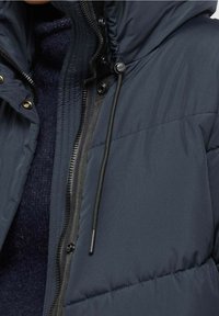 Marinblå pufferjacka med huva, med dragkedja och tryckknappsstängning, strukturerad tyg med horisontella sömmar och snörning vid halsen.