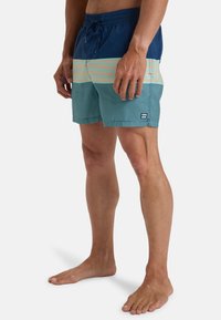 Pánske plavky vyrobené z ľahkej tkaniny, námornícka modrá s horizontálnymi zelenými a broskyňovými pruhmi, s pásom na sťahovanie a detailom loga.