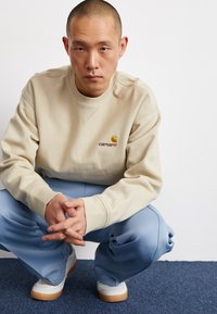 Beige sweatshirt met Carhartt-logo, ronde hals, geribbelde manchetten; gecombineerd met losse blauwe broek en witte sneakers met gumachtige zolen. Hurkende pose.