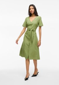 Femme en robe midi olive boutonnière avec ceinture à la taille et manches courtes, portant des mules à talons aiguille noires, se tenant sur fond blanc.