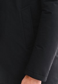 Czarna kurtka puchowa z gładką, wyściełaną teksturą, z boczną kieszenią i lekko zwężanym mankietem.