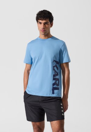 Man draagt een lichtblauw T-shirt met de tekst "KARL" aan de zijkant en een zwarte korte broek met kleine witte gezichtspictogrammen op het been.